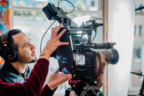 A person wearing a red long-sleeved shirt operates a video camera, while another person observes the scene being filmed